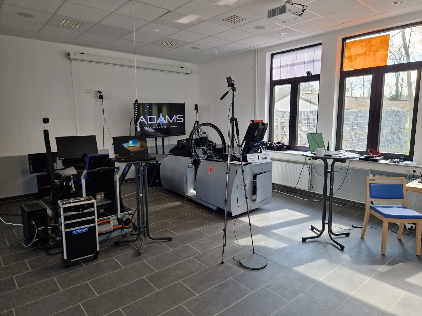 The image shows the Experiment setup of the XR Emergency Training for Military Fast Jets. It shows a flight simulator, infront of a screen, with different cameras and monitors around it.